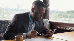 Black businessman looks at smartphone over top of his paperwork. Hot cup of coffee on the table. He wears shirt and suit jacket.