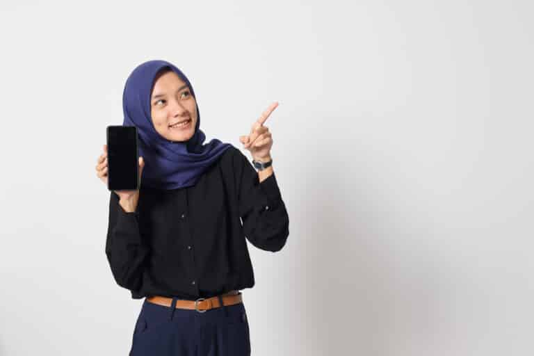Portrait of excited Asian woman in button-up shirt and hijab, showing blank screen mobile phone mockup while pointing and presenting product. Isolated image on white background.