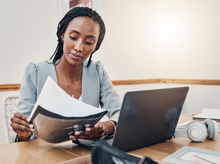 A woman in a blazer sits at a desk reviewing paperwork in front of her laptop.