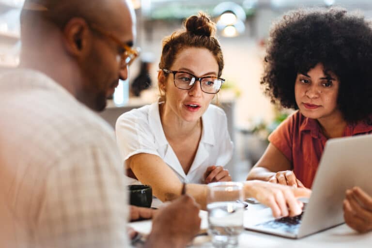 Two women and a man in a business setting gather around a laptop that one woman points at.